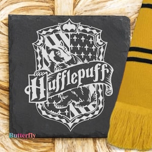Könnte beinhalten: Ein quadratischer, dunkelgrauer Schieferuntersetzer mit dem Hufflepuff-Hauswappen aus der Harry-Potter-Reihe. Das Wappen ist weiß und enthält einen Dachs, den Hausnamen und andere dekorative Elemente. Ein gelb-schwarzer Schal ist teilweise sichtbar.