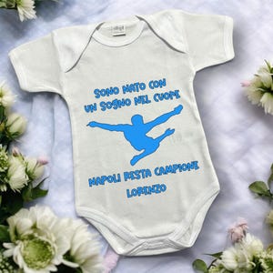 Può includere: Body da bambino bianco con testo blu e sagoma blu di una persona. Il testo recita "SONO NATO CON UN SOGNO NEL CUORE NAPOLI RESTA CAMPIONE LORENZO."