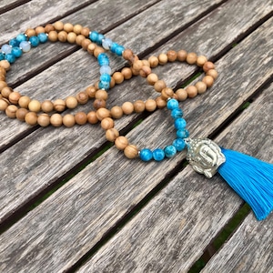 Könnte beinhalten: Eine lange Perlenkette mit runden, hellbraunen Holzperlen, die mit leuchtend blauen und weißen marmorierten Perlen durchsetzt sind. Die Kette hat einen silberfarbenen Buddha-Kopf-Anhänger aus Metall und eine leuchtend blaue Stoffquaste.