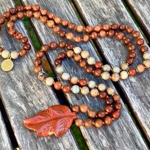 Può includere: Una collana artigianale con perline di legno nei toni del marrone e beige, infilate su un cordino. Include un ciondolo circolare dorato e un ciondolo a forma di foglia intagliata. Le perline variano per dimensione e colore, creando un'estetica naturale e terrosa.