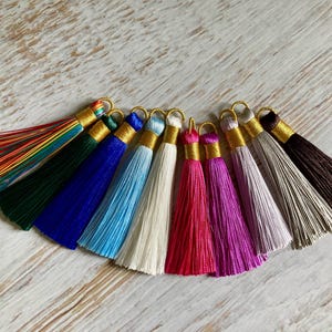 Puede incluir: Una colección de borlas coloridas en varios tonos, incluyendo arcoíris, verde, azul, celeste, blanco, rojo, rosa, morado, gris y marrón. Cada borla tiene una tapa y un lazo dorados para colgar.