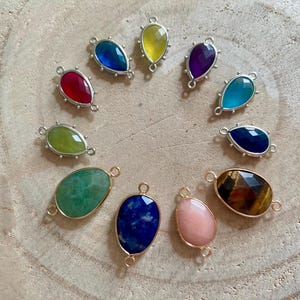 Könnte beinhalten: Eine Sammlung von elf tropfenförmigen Schmuckverbindern mit verschiedenen Edelsteinen und Glassteinen in Farben wie Rot, Blau, Gelb, Lila, Grün und Braun. Jedes Stück ist in eine metallische Fassung mit zwei kleinen Ösen eingefasst.