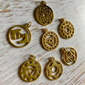 Op de afbeelding: Een verzameling van zeven goudkleurige hangers met diverse geometrische en symbolische ontwerpen. De ronde hangers hebben ingewikkelde patronen zoals sterren, driehoeken en bloemmotieven. Elke hanger heeft een kleine lus om aan een ketting of koord te bevestigen.