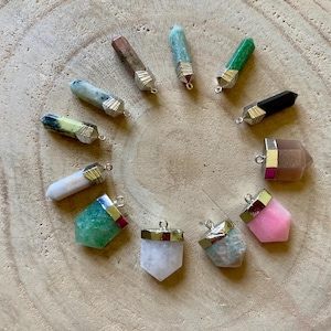 Puede incluir: Una colección de doce colgantes de cristal con forma de punta dispuestos en círculo. Cada colgante presenta una piedra preciosa pulida en varios colores, incluyendo rosa, verde, negro, blanco y marrón, con una tapa de metal plateado y una anilla.