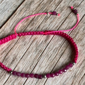 Può includere: Un vivace braccialetto in macramè rosa con una fila di perline rosa intenso sfaccettate. Il braccialetto è dotato di una chiusura a nodo scorrevole ed è impreziosito da piccole perline argentate. Il braccialetto regolabile è esposto su una superficie di legno chiaro.