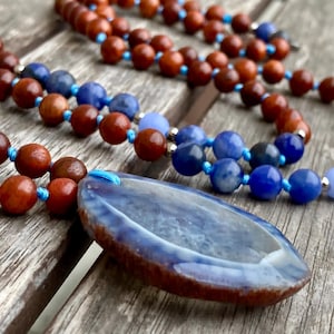Pode incluir: Um colar de contas com contas redondas de madeira castanha e contas de pedra azul, enfiadas num cordão azul brilhante. O colar inclui um pendente de ágata grande, polido e oval, em tons de azul e castanho, com uma superfície lisa e translúcida.