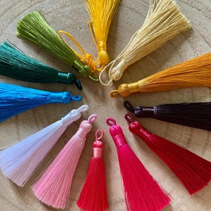 Könnte beinhalten: Eine Sammlung von zwölf dekorativen Quasten in kreisförmiger Anordnung, die ein lebendiges Farbspektrum in Gelb, Creme, Orange, Schwarz, Rot, Magenta, Pink, Weiß, Blau, Türkis und Grün zeigen, jede mit einer kleinen Schlaufe an der Oberseite.