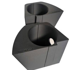 Op de afbeelding: Twee matte zwarte, geometrische kandelaars. Elke houder heeft een cirkelvormige opening en een platte basis. Het woord "CHPARIS" is zichtbaar op één houder. Een klein, wit, cirkelvormig object bevindt zich in een van de houders.