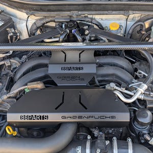 Peut inclure: Un compartiment moteur avec des couvercles en métal noir. Les couvercles portent les inscriptions "86PARTS" et "GREENFUCHS" en blanc. Les composants du moteur sont visibles, y compris des tubes et d'autres pièces mécaniques. Le capot de la voiture est ouvert.