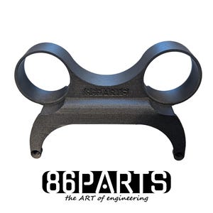 Può includere: Un componente automobilistico nero opaco stampato in 3D con due aperture circolari nella parte superiore e una base curva con due rebbi rivolti verso il basso. Il testo 86PARTS è impresso sul ponte centrale. Sotto l'oggetto, il logo 86PARTS the ART of engineering è visualizzato in nero.
