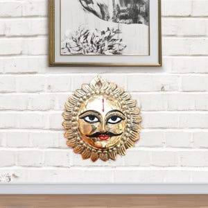 Könnte beinhalten: Eine goldfarbene Metallsonne mit einem Gesicht, das mit schwarzen Augen, einem roten Mund und einem weißen vertikalen Strich auf der Stirn bemalt ist. Die Sonne ist von einem dekorativen, floralen Design umgeben.