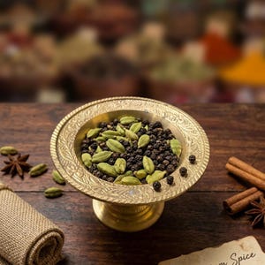 May include: A gold-toned metal pedestal bowl with an ornate engraved rim, filled with a mixture of green cardamom pods and black peppercorns. The bowl sits on a dark wooden surface alongside scattered star anise, cinnamon sticks, and burlap.