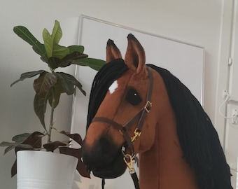 Può includere: Un cavallo a dondolo marrone con una criniera nera e una macchia bianca sulla fronte. Ha una briglia marrone con finiture color oro e una redine nera. Il cavallo è montato su un bastone di legno.