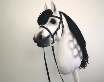 May include: A white and gray hobby horse with a black mane and bridle. The toy horse has a wooden stick for a handle and a black strap. The horse has a painted face and is designed to look like a real horse.