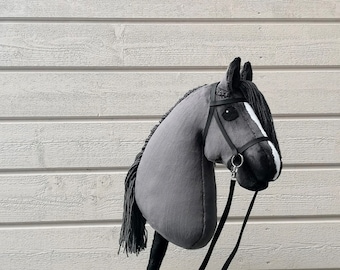May include: A gray hobby horse with a black mane and tail, featuring a black bridle and reins. The toy horse is mounted on a light-colored wooden stick.