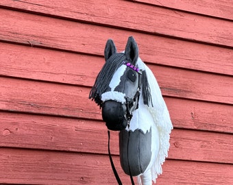 May include: A gray and white hobby horse with a black bridle and purple beads on the mane. The horse is standing on a wooden stick and has a black strap.
