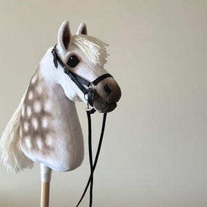 May include: A soft toy hobby horse with a white body and brown spots. It features a white mane and tail, a black bridle, and a black lead rope. The horse is attached to a wooden stick.