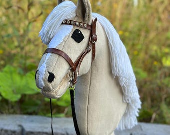 May include: A beige hobby horse with a white mane and tail. The horse has a brown bridle with silver-colored hardware and a black lead rope. The horse's head is made of soft fabric and has black eyes.
