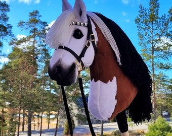 Puede incluir: Una cabeza de caballo marrón y blanca con una crin y cola negras. El caballo lleva una cabezada de cuero negra con herrajes dorados y una cuerda de plomo negra. El caballo está de pie en un campo con árboles y un cielo azul de fondo.