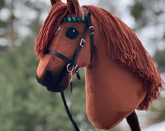 Puede incluir: Una cabeza de caballo de peluche marrón con una brida negra y una diadema de cuentas turquesa. La cabeza del caballo está unida a un palo de madera.