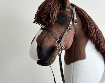 Puede incluir: Una cabeza de caballo de peluche marrón y blanco con una brida de cuero marrón y riendas negras. El caballo tiene una crin y una cola marrón.