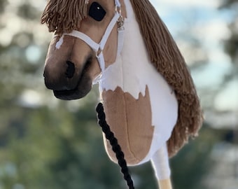 Op de afbeelding: Een bruin en wit pinto-paardenhoofd hobbypaard met een wit halster en zwarte teugels. Het paard heeft een bruine manen en staart.