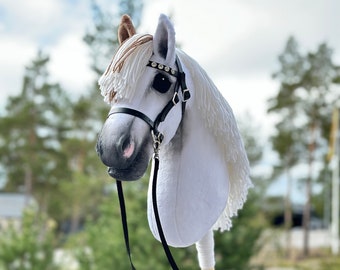 Puede incluir: Cabeza de caballo de peluche blanca con una brida y riendas negras. El caballo tiene una crin y una cola marrones. El caballo está montado en un palo de madera.