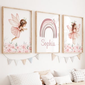 May include: Three framed prints in a pink and white colour scheme. The prints feature a fairy with wings, a rainbow with the name "Sophia" written below it, and another fairy with wings. The prints are hanging on a white wall above a wooden shelf with storage bins.