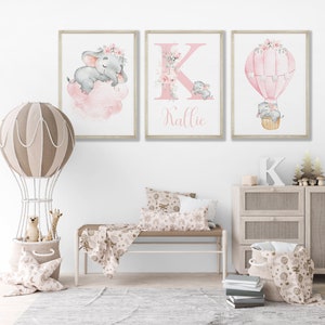 May include: Three watercolor prints featuring a pink and gray elephant, a pink letter K with floral accents, and a pink hot air balloon with floral accents. The prints are framed in a light wood frame and have the name "Kallie" written below the letter K.