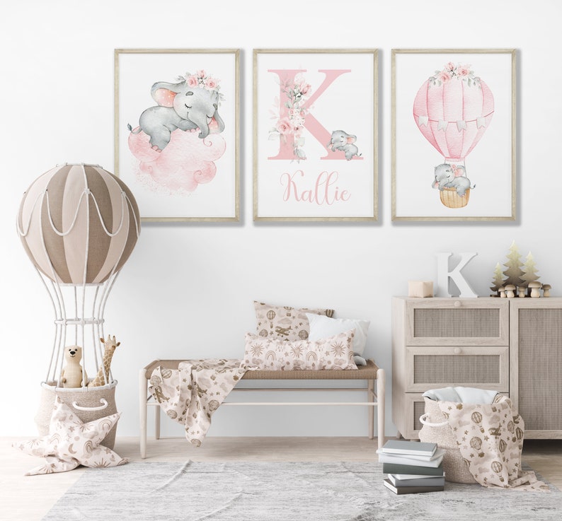 May include: Three watercolor prints featuring a pink and gray elephant, a pink letter K with floral accents, and a pink hot air balloon with floral accents. The prints are framed in a light wood frame and have the name "Kallie" written below the letter K.