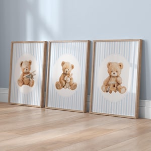May include: Three framed prints featuring watercolor illustrations of teddy bears. Each bear is depicted in a different pose, holding a toy airplane, a stuffed bunny, and a wooden train. The background has a blue and white striped pattern.