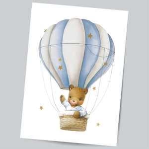 Op de afbeelding: Een aquarel-illustratie van een blauw-wit gestreepte luchtballon met een bruine teddybeer die een blauw-wit gestreept shirt draagt ​​erin. De ballon is omringd door kleine gouden sterren.