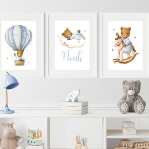 Op de afbeelding: Drie ingelijste prints met een blauwe en witte luchtballon, een teddybeer die op een wolk slaapt en een teddybeer die een schommelpaard berijdt. De middelste print heeft de naam Noah in blauw.