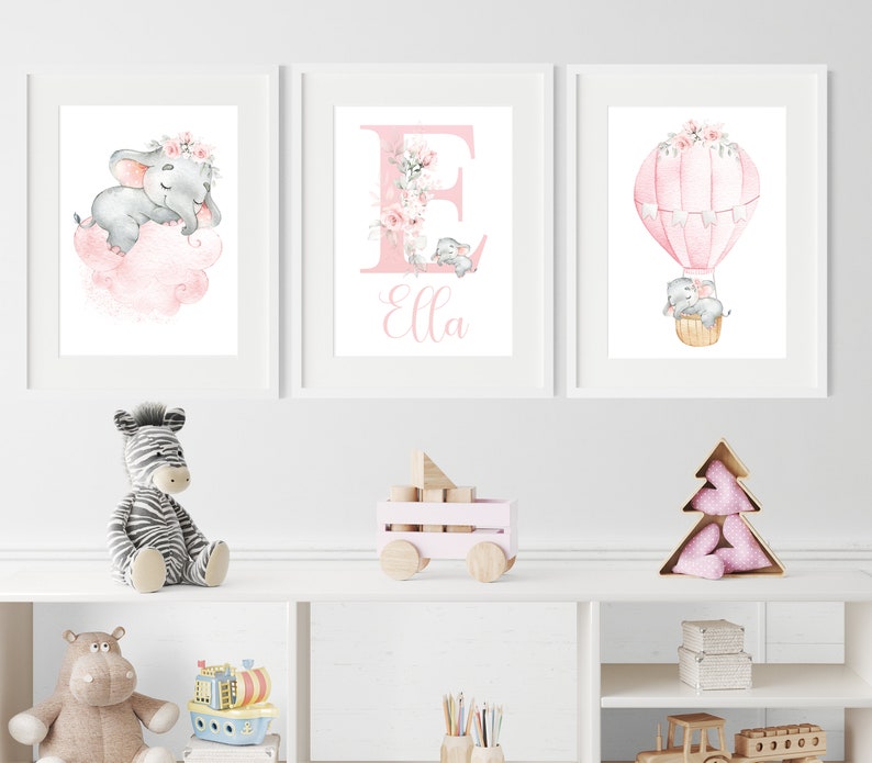 May include: Three framed prints with a pink and gray color scheme. The first print features a pink cloud with a sleeping elephant wearing a flower crown. The second print features a pink letter E with a floral design and the name Ella. The third print features a hot air balloon with two elephants inside.