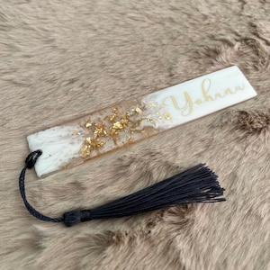 Könnte beinhalten: Weißes Lesezeichen mit goldenen Flocken und dem Namen "Yohana" in goldenen Buchstaben. Das Lesezeichen hat einen schwarzen Quasten.