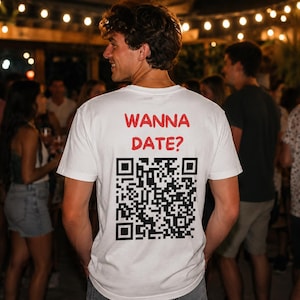 以下が含まれることがあります： 背面に赤い文字で「WANNA DATE?」とプリントされ、その下に大きな白黒のQRコードが配置された白い半袖Tシャツ。後ろ姿で撮影されており、白い生地の上にグラフィックデザインがはっきりと表示されています。