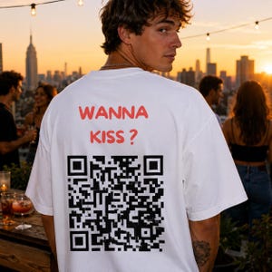 以下が含まれることがあります： 背面に赤い文字で「WANNA KISS?」とプリントされ、その下に大きな正方形の白黒QRコードが配置された白い半袖Tシャツ。デザインはクルーネックの衣服の背中上部中央に配置されています。
