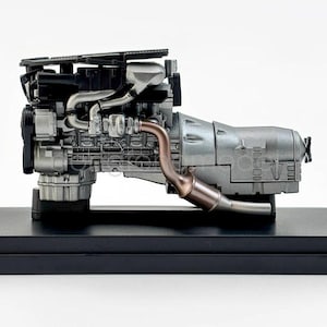 May include: A detailed model engine, predominantly silver and black, with copper-coloured exhaust pipes. The engine is mounted on a black rectangular base, displaying its intricate mechanical components.