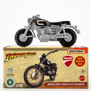May include: A silver and black Matchbox toy motorcycle, an Indiana Jones themed Ducati Scrambler, sits on its cardboard box. The box features the Matchbox logo, Ducati logo, and "NEW NOUVEAU 2024" text. The toy is designed for ages 3+.