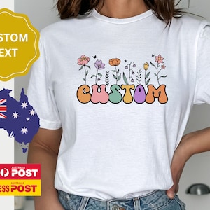 Op de afbeelding: Wit T-shirt met een kleurrijke afbeelding van het woord "CUSTOM" in een retro-lettertype met bloemen die uit de letters groeien.