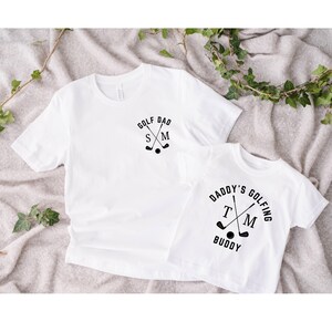 May include: Two white t-shirts with black text and graphics. The larger shirt says "Golf Dad" with crossed golf clubs and the initials "S M". The smaller shirt says "Daddy's Golfing Buddy" with crossed golf clubs and the initials "T M".