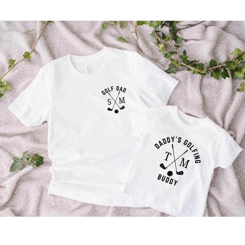 May include: Two white t-shirts with black text and graphics. The larger shirt says "Golf Dad" with crossed golf clubs and the initials "S M". The smaller shirt says "Daddy's Golfing Buddy" with crossed golf clubs and the initials "T M".