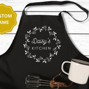 May include: Black kitchen apron with a white floral wreath design and the text "Daisy's Kitchen".