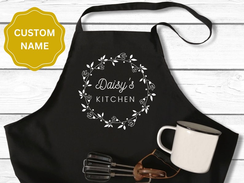 May include: Black kitchen apron with a white floral wreath design and the text "Daisy's Kitchen".