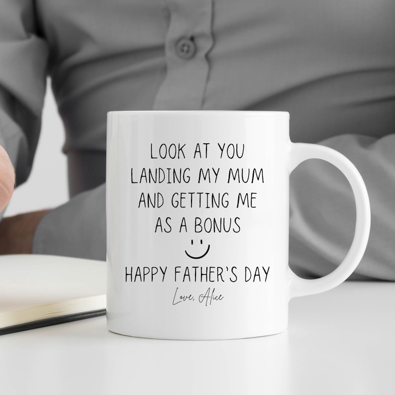 May include: White ceramic mug with a black printed message: "LOOK AT YOU LANDING MY MUM AND GETTING ME AS A BONUS" with a smiley face and "HAPPY FATHER'S DAY Love, Alice". The mug has a curved handle.