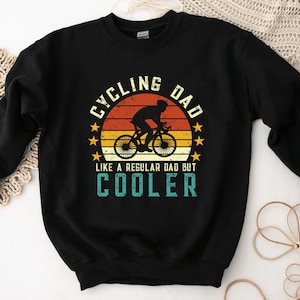 Pu&ograve; includere: Felpa nera con una grafica in stile vintage che dice "Cycling Dad Like a Regular Dad But Cooler". La grafica presenta una silhouette di una persona che guida una bicicletta davanti a un tramonto.
