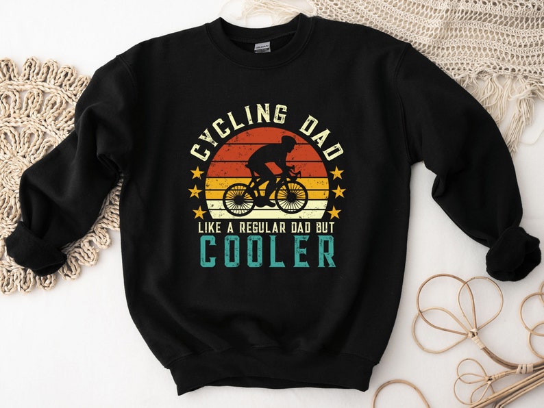 Pu&ograve; includere: Felpa nera con una grafica in stile vintage che dice "Cycling Dad Like a Regular Dad But Cooler". La grafica presenta una silhouette di una persona che guida una bicicletta davanti a un tramonto.