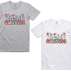 Op de afbeelding: Twee T-shirts met een bloemenontwerp en het woord "CUSTOM" in kleurrijke bubbelletters. De shirts zijn grijs en wit.