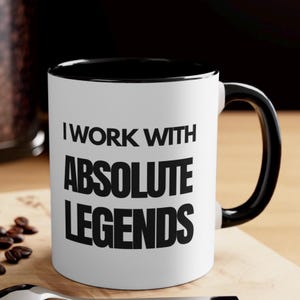 May include: White ceramic coffee mug with a black rim and handle. The mug has black text that reads "I work with absolute legends".