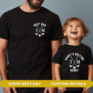 May include: Two black t-shirts with a golf theme. The larger shirt says "Golf Dad" with crossed golf clubs and initials "S M". The smaller shirt says "Daddy's Golfing Buddy" with crossed golf clubs and initials "T M".